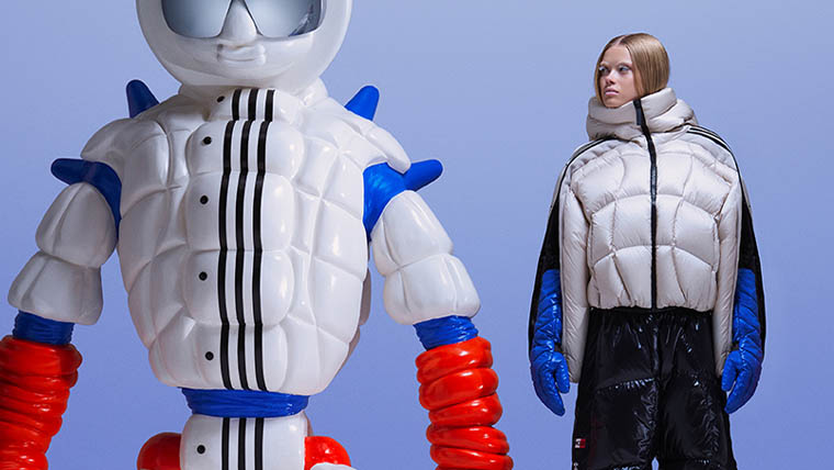 Eine blonde Frau trägt eine silberfarbene adidas x Moncler Daunenjacke mit blauen Handschuhen und steht neben einer weißen Figur (Foto)