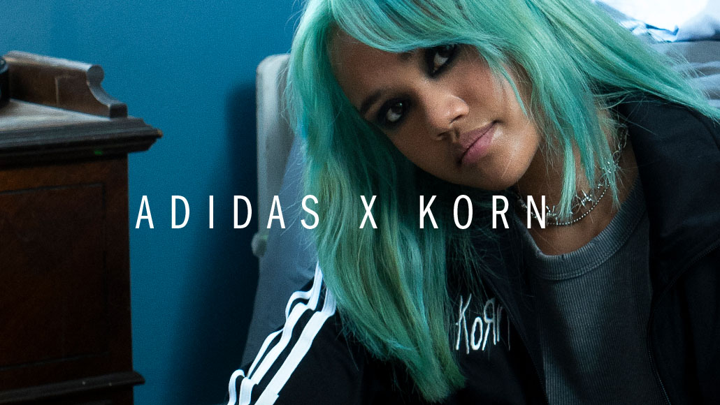 A woman with turquoise hair looks into the camera and wears a black adidas x Korn jacket (Photo)