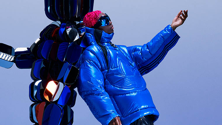 A person with red hear wears sunglasses and a blue adidas x Moncler down jacket. They lean against a figure made of blue cubicles (Photo)