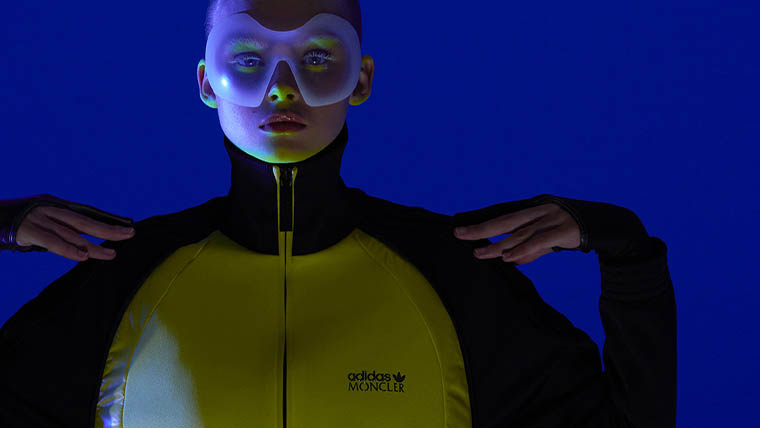 A woman with yellow eye shadow wears a yellow adidas x Moncler jacket and stands in front of a blue background (Photo)