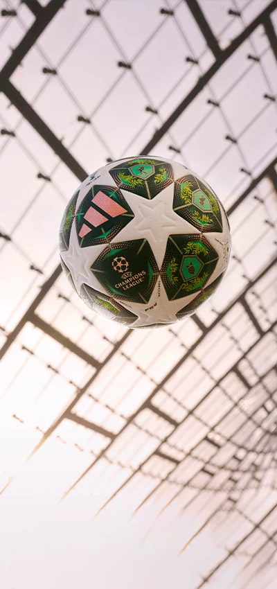 Dieses Bild zeigt die Innenansicht eines Stadions, das wie ein Fußballstadion aussieht, mit Wänden und Decke aus perforiertem Metallgeflecht. Im Vordergrund ist ein gemusterter Ball mit den Sternen der UEFA Champions League zu sehen.