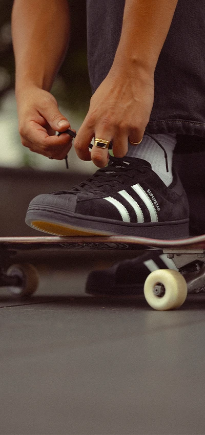 Ein Bild einer Person, die ein Paar schwarze Adidas-Sneaker trägt, auf einem Skateboard steht und die Schnürsenkel bindet.