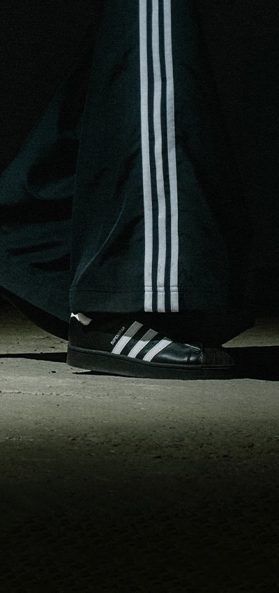 Auf dem Foto sind das Bein und der Fuß einer Person zu sehen, die Adidas-Sportbekleidung und Adidas-Schuhe trägt, beide in Schwarz mit weißen Streifen.