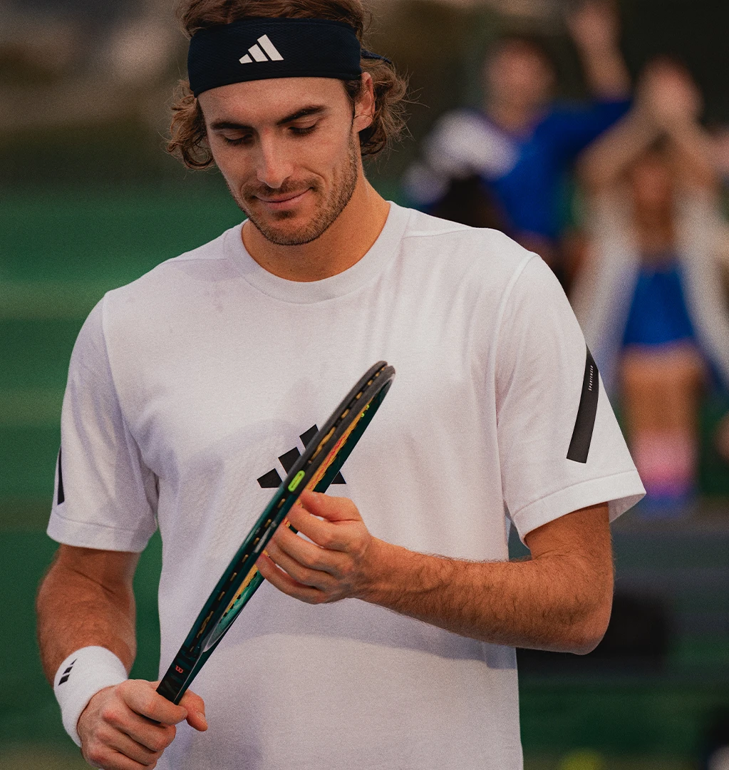 Das Bild zeigt einen Tennisspieler, der seinen Schläger hält und auf ihn herabblickt. Er trägt legere Sportkleidung, bestehend aus Shorts mit einem Adidas-Logo, einem passenden ärmellosen Oberteil und einem Stirnband.