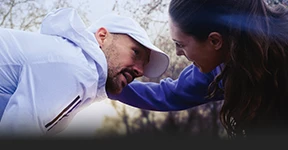 Dies ist ein Foto von zwei Personen. Links ist ein Mann mit dunklem Haar, der einen weißen Adidas-Trainingsanzug und eine weiße Kappe trägt. Rechts ist eine Frau mit langen Haaren zu einem Pferdeschwanz, sie lehnt sich leicht nach vorne zu dem Mann, gestikuliert vielleicht oder spricht mit ihm. Der Hintergrund ist verschwommen, aber es sieht aus wie eine Außenkulisse bei Tageslicht mit natürlichem Licht.