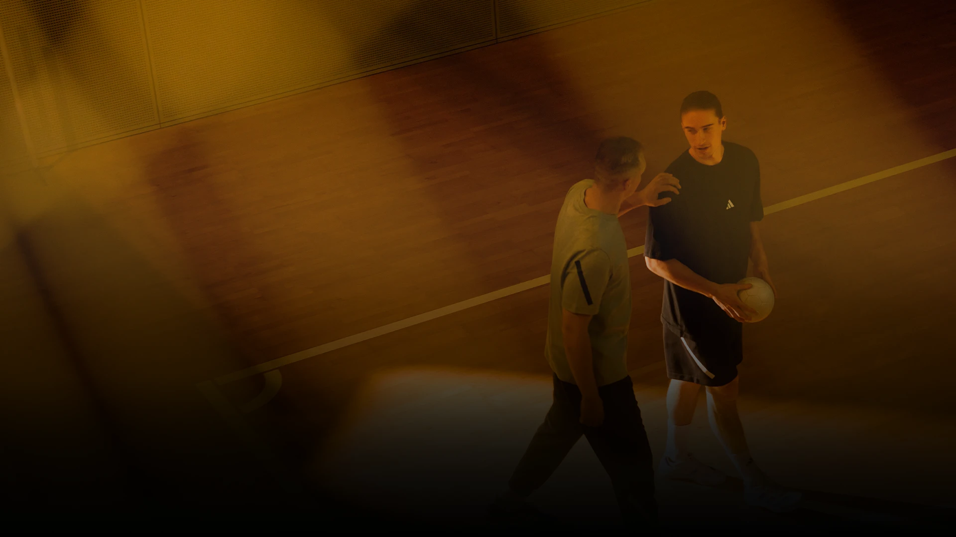 Auf diesem Foto sind zwei Männer zu sehen, die auf einer Fläche stehen, die wie ein Basketballfeld aussieht. Einer von ihnen hält einen Sportball, während der andere in ein Gespräch vertieft oder anhand der Gesten und Körpersprache der ersten Person interagiert.