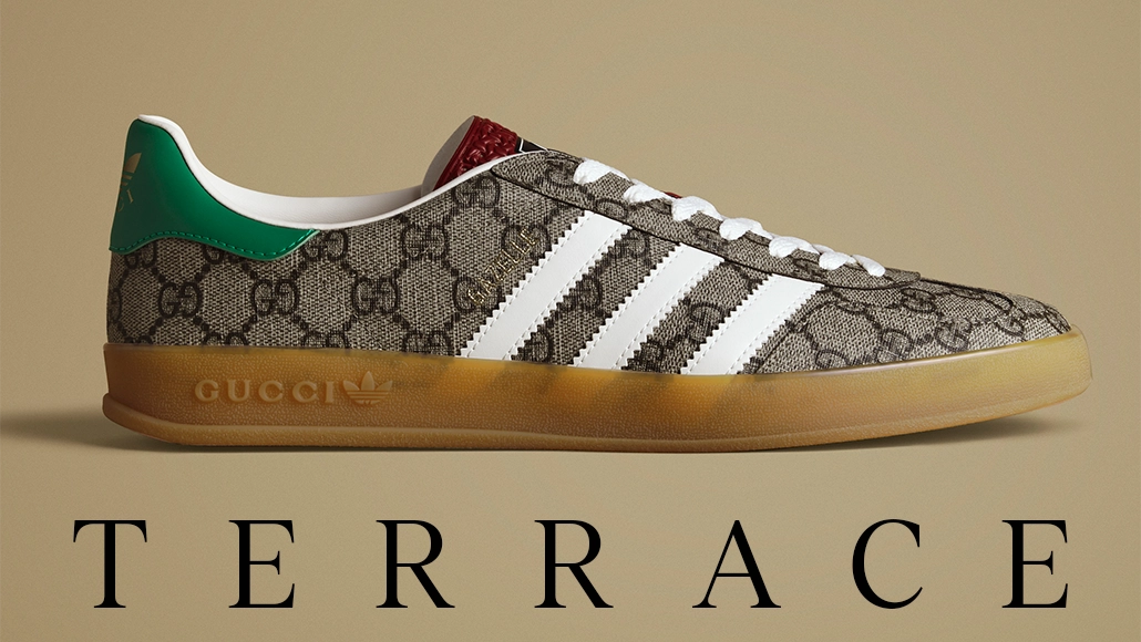 Das Bild zeigt ein Paar Adidas-Schuhe im Sneaker-Design. Vor den Schuhen steht das Wort „Terrace“ in grafischer Schrift. Der Markenname „GUCCI“ überlappt eine Seite des Schuhs, was darauf hindeutet, dass es sich um eine Zusammenarbeit mit Gucci handeln könnte. In der unteren rechten Ecke befindet sich ein kleines Etikett mit dem Adidas-Logo, das darauf hinweist, dass es sich tatsächlich um Adidas-Schuhe handelt. Der Hintergrund ist neutral und legt den Fokus ganz auf die Schuhe und den Text.