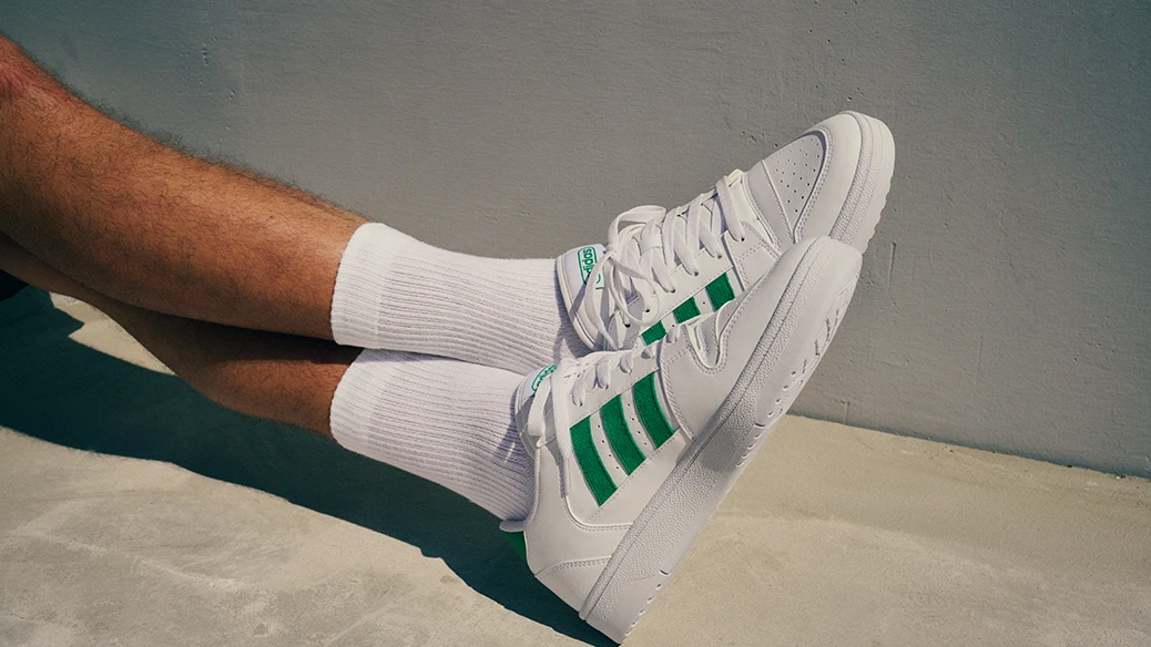 The image shows a person's legs and feet with adidas shoes on them, positioned against a pale surface.