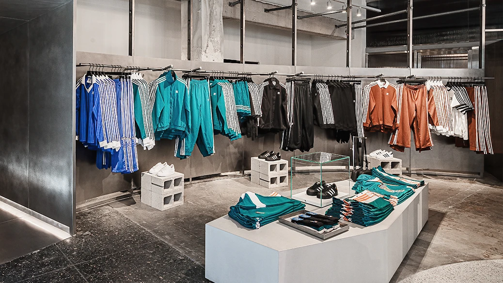 The image shows an interior view of an adidas store. There is a variety of jackets and other sportswear displayed on the walls The color scheme of the items primarily consists of dark and light colors with striped patterns.