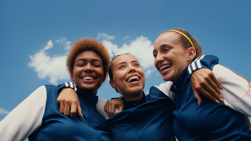 The image shows a group of female athletes, who appear to be celebrating or preparing for an event. They are dressed in sports attire and look like they might be teammates, wearing tracksuits with adidas brand logos visible on the sleeves.