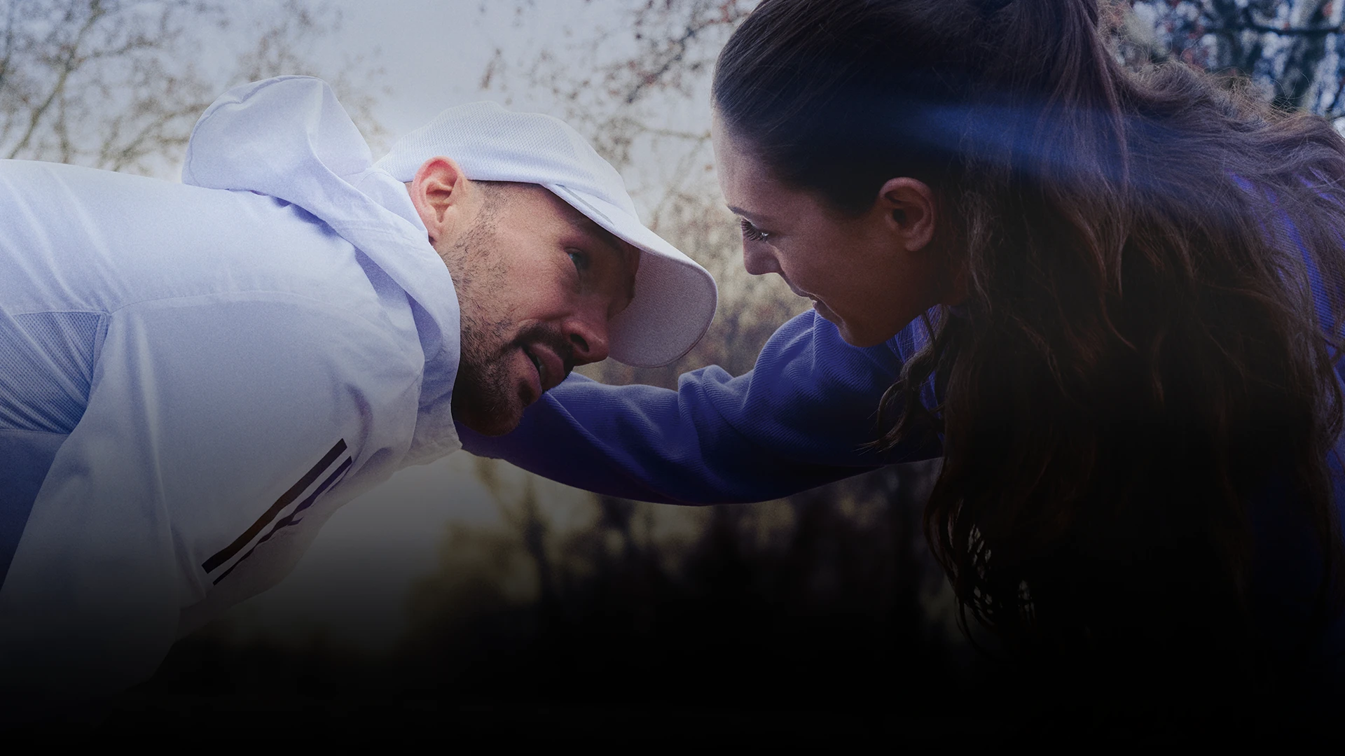 This is a photo of two people. On the left, there is a man with dark hair who is wearing a white adidas tracksuit anda qhite cap. On the right, there's a woman with long hair in a ponytail, she is leaning slightly forward towards the man, possibly gesturing or speaking to him. The background is blurred, but it looks like an outdoor setting during the daytime with natural light.