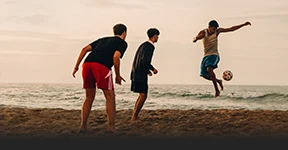 This photo shows people playing soccer on a beach at either sunrise or sunset due to the warm glow and color saturation. The image captures a moment of motion and activity, possibly from a social gathering or event involving sports. 