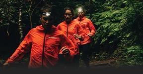 The image shows three individuals wearing orange jackets, likely from a team or company, and they appear to be on a hike or a run in a forested area. They all have headlamp attachments, suggesting they are either out after dark or that it's twilight or overcast.