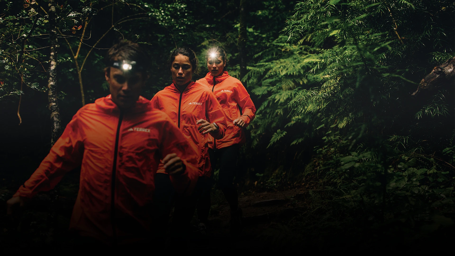 The image shows three individuals wearing orange jackets, likely from a team or company, and they appear to be on a hike or a run in a forested area. They all have headlamp attachments, suggesting they are either out after dark or that it's twilight or overcast.