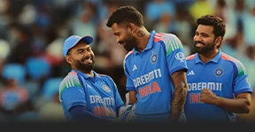 The image shows three men posing towards the camera. They are wearing cricket uniforms with logos indicating they are part of the Indian cricket team. One player is smiling directly at the camera while the other two are laughing and looking away from it.