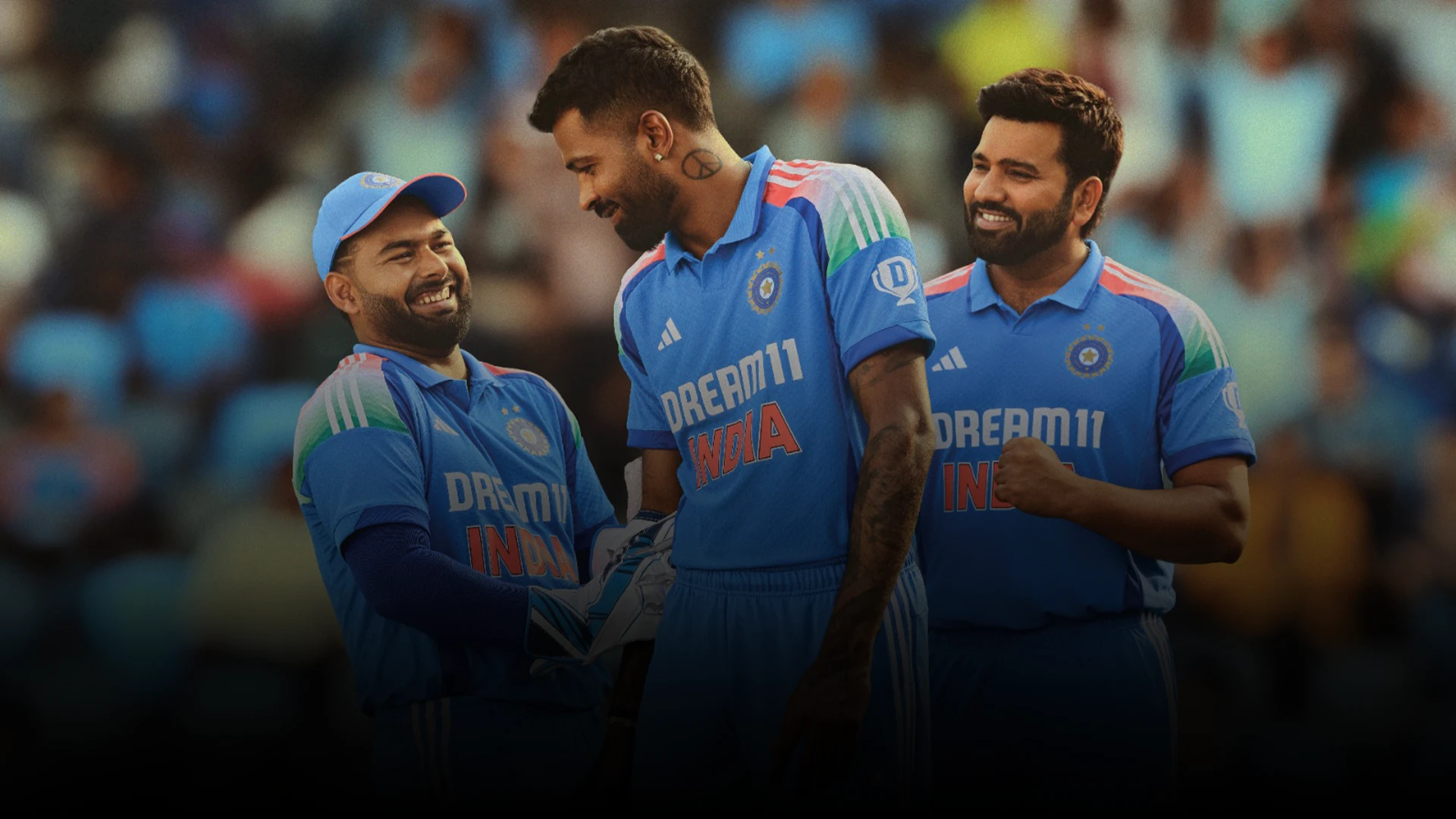 The image shows three men posing towards the camera. They are wearing cricket uniforms with logos indicating they are part of the Indian cricket team. One player is smiling directly at the camera while the other two are laughing and looking away from it.