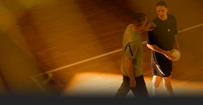 This photo shows two men standing on what appears to be a basketball court, one of whom is holding a sports ball, while the other seems engaged in conversation or interaction with the first person's gestures and body language.