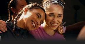 The image is a photograph capturing two individuals embracing. The person on the left seems to be smiling and is wearing some type of athletic clothing, while the person on the right has long hair, a smile, and is also in an athletic outfit.