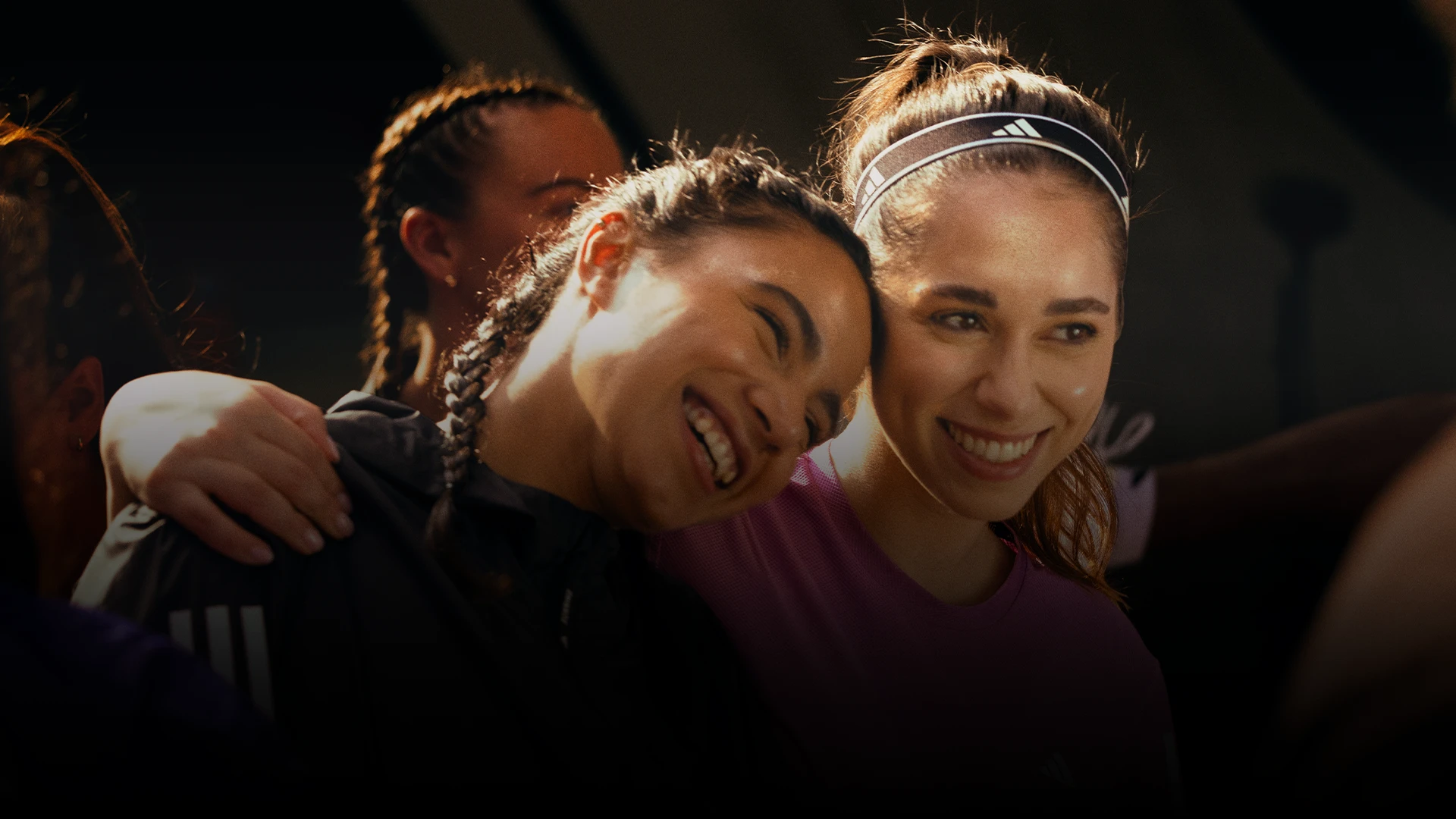 The image is a photograph capturing two individuals embracing. The person on the left seems to be smiling and is wearing some type of athletic clothing, while the person on the right has long hair, a smile, and is also in an athletic outfit.