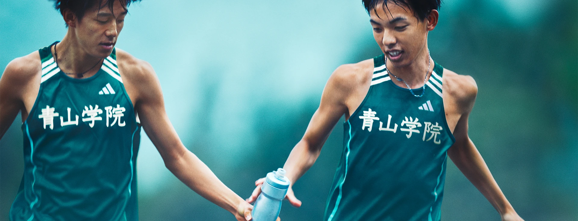 This is a color photograph featuring two male athletes, likely in mid-stride running track races. They are wearing light green sleeveless tops with the logo of their athletic club, possibly Japan given the text and design on the uniforms which include characters that are also present in Japanese.