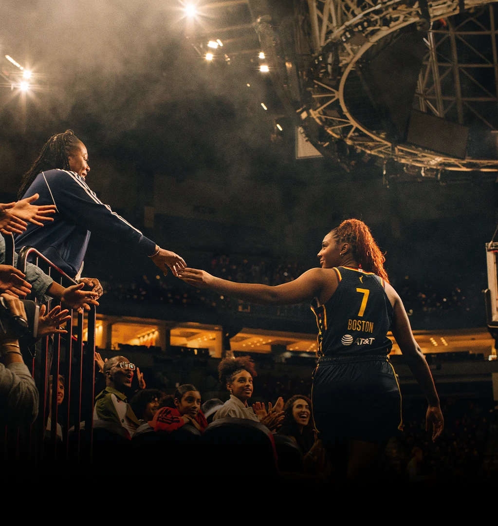 The image shows a sporting event. In the foreground, there is a crowd of people watching what seems to be a women's basketball game. A player from one team can be seen shaking hands with or receiving a hand from someone, possibly a teammate or coach, mid-action. 