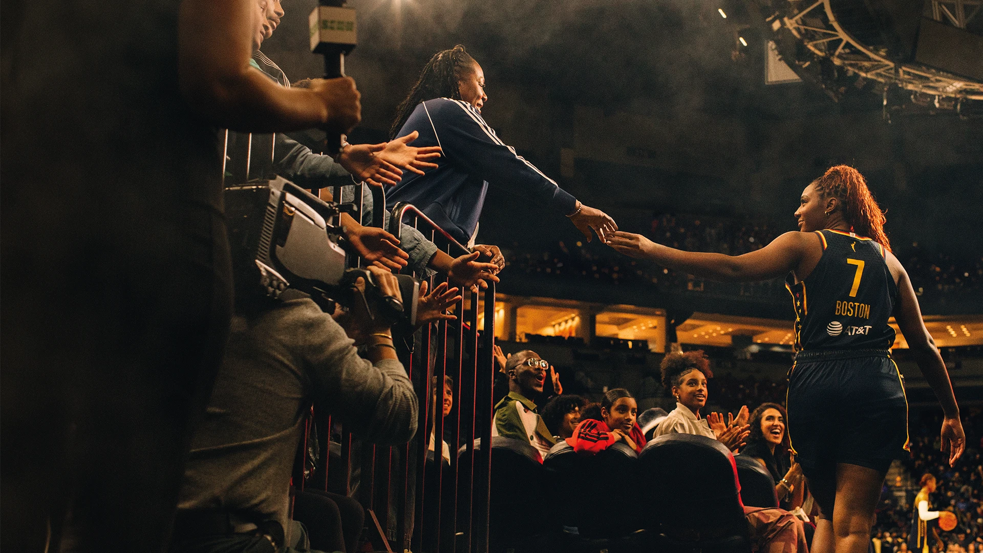 The image shows a sporting event. In the foreground, there is a crowd of people watching what seems to be a women's basketball game. A player from one team can be seen shaking hands with or receiving a hand from someone, possibly a teammate or coach, mid-action. 