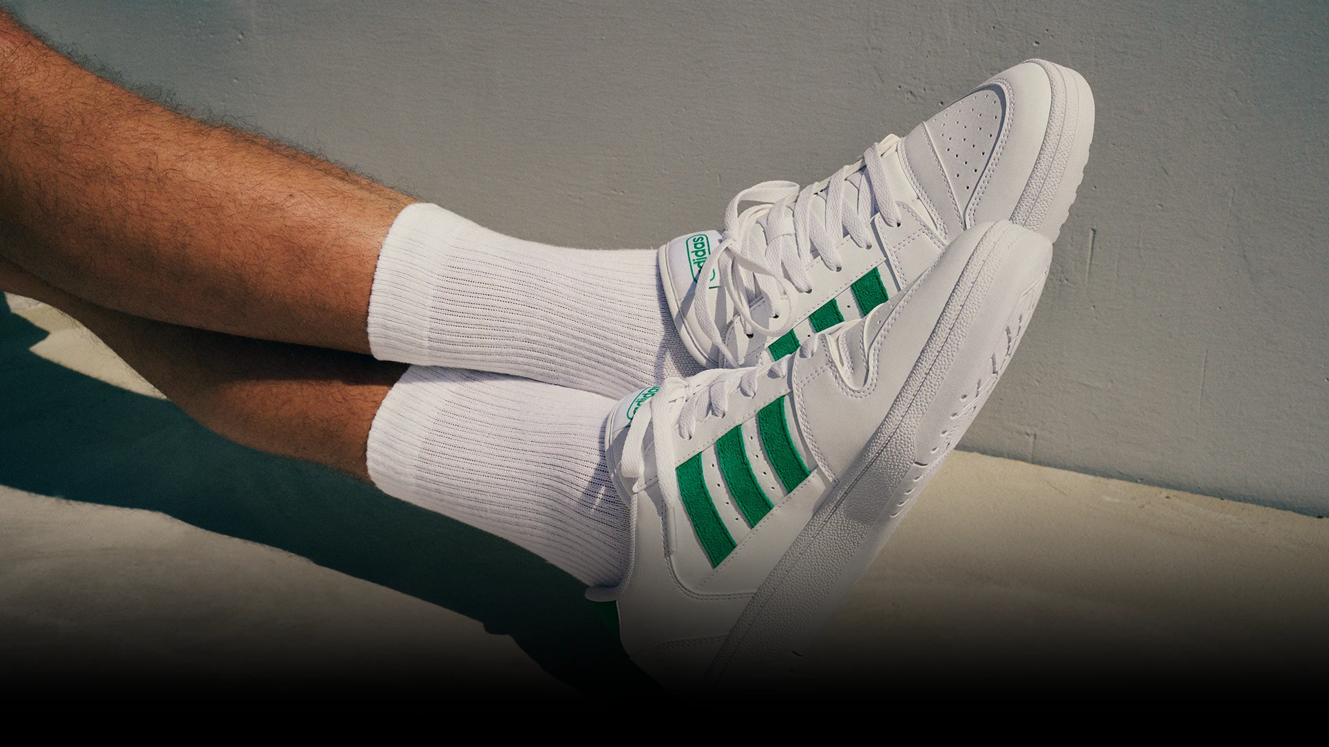 The image shows a pair of white sneakers with green accents, possibly the Adidas ZX500 model or another similar design that resembles this colorway. The sneakers are placed next to what appears to be a leg wearing long socks that match the shoes, with a hint of sports apparel above the ankle. There is no person fully visible in the photo.