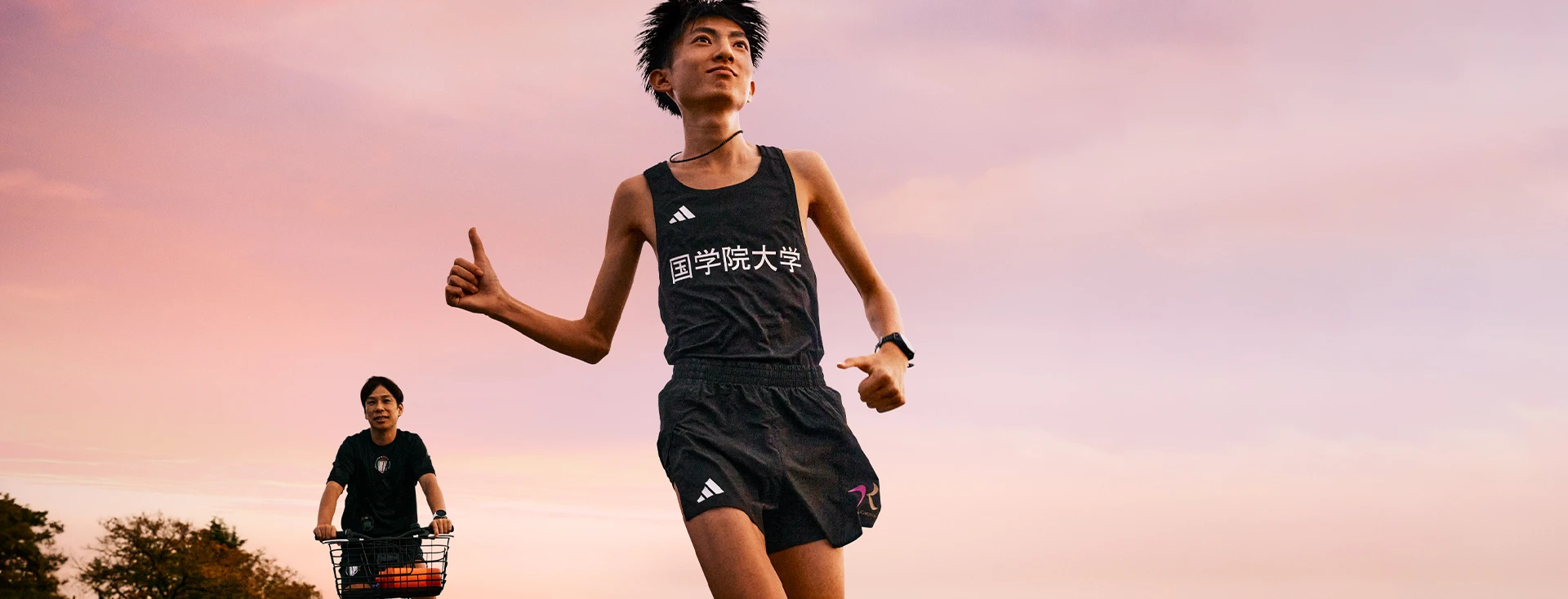 This is an image of a person jogging outdoors. The background shows a clear sky during what seems to be either dawn or dusk, given the warm colors suggesting either sunrise or sunset. There's also another individual on a bicycle in the background, but they are too far away to discern any specific details about them. The jogger is wearing sports attire and seems to be enjoying their run.