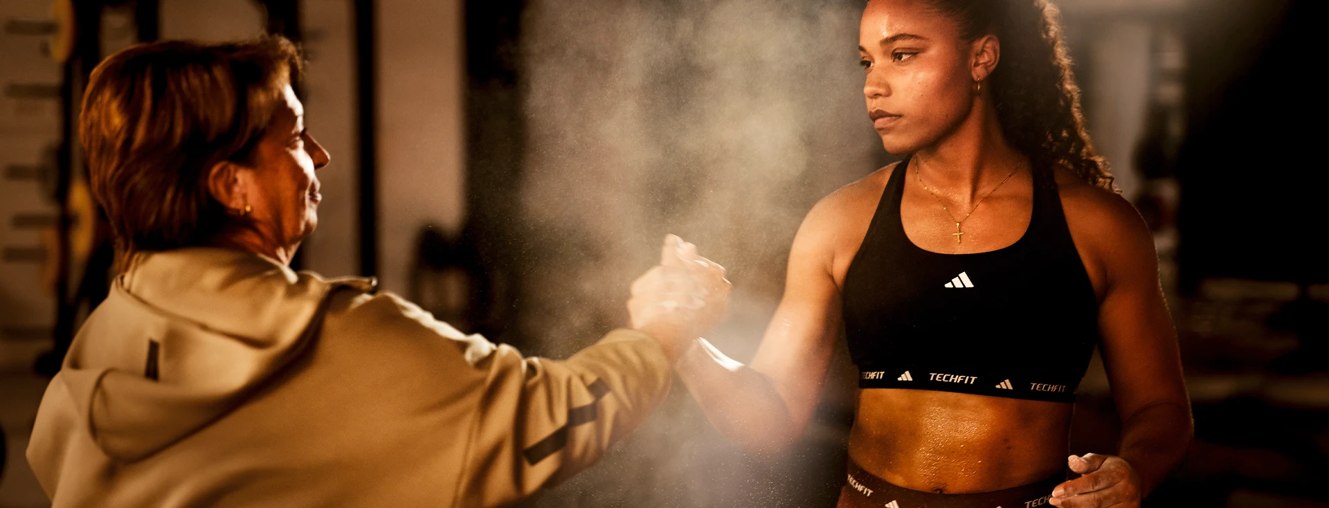 In the image, you can see two women who appear to be interacting with each other in a gym or workout facility. They are surrounded by weights and various exercise equipment, which suggests that they may be engaged in an exercise-related discussion or demonstration.