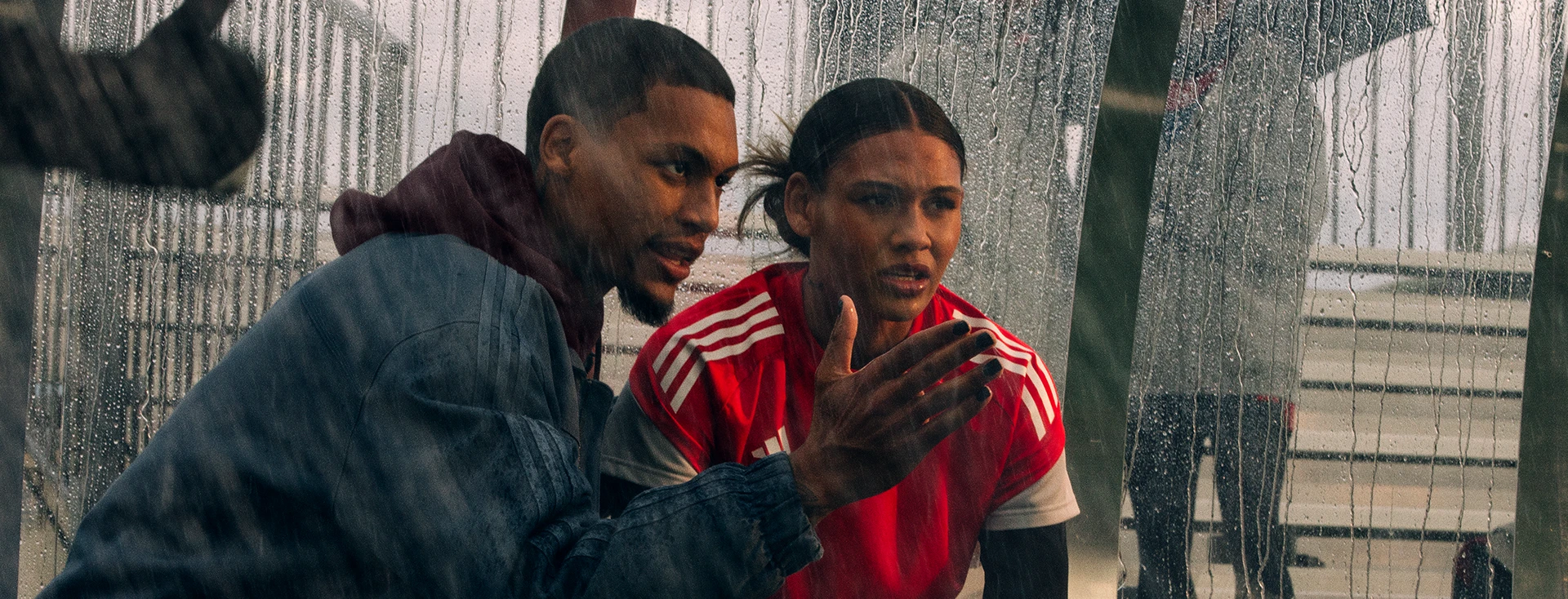 This is a photograph of two individuals. They both seem to be engaged in a conversation or observing something together that isn't visible in the photo. Both people are dressed in sport outfits and are standing near what looks like a fence of a stadium.