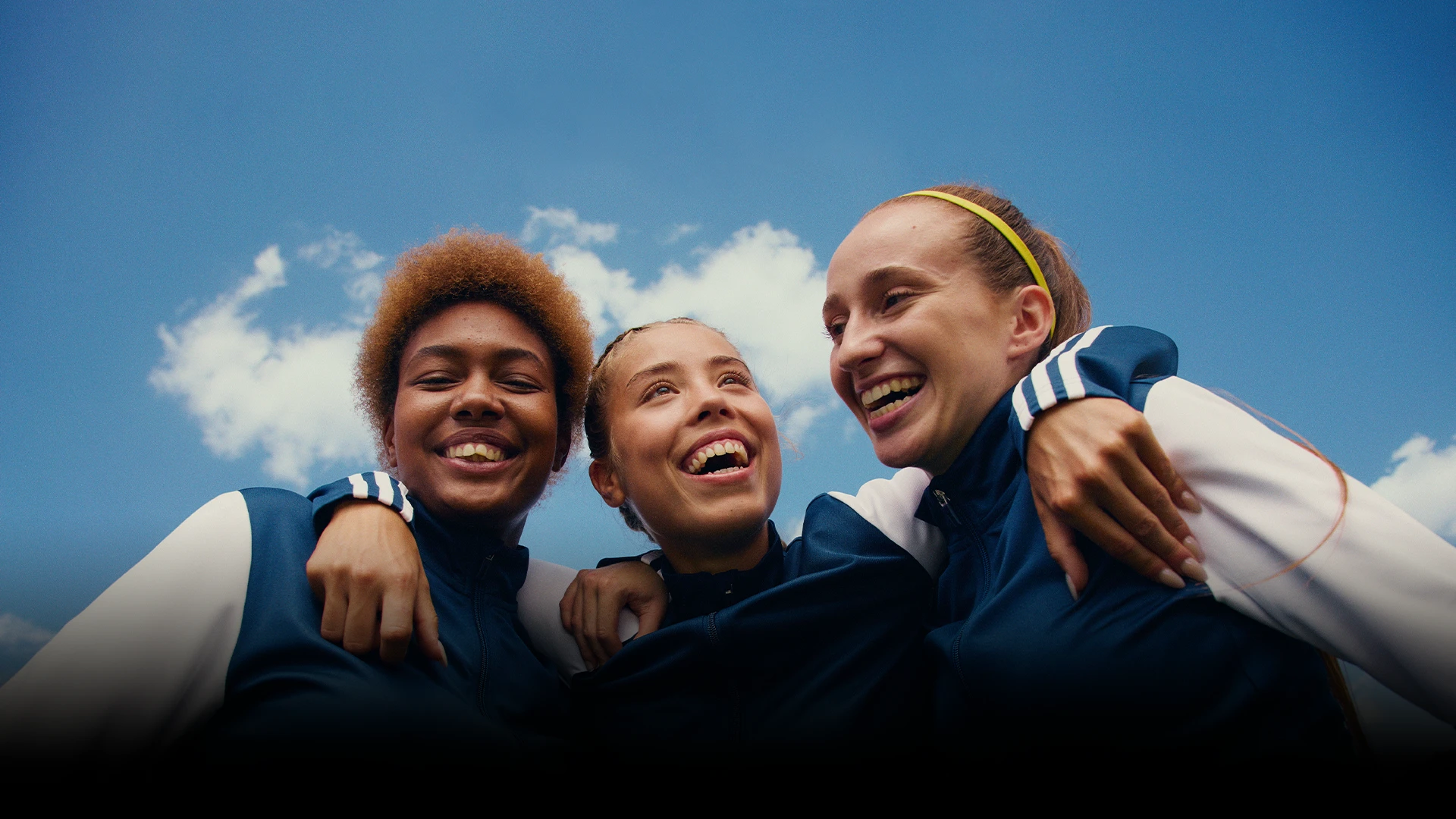 The image shows a group of female athletes, who appear to be celebrating or preparing for an event. They are dressed in sports attire and look like they might be teammates, wearing tracksuits with adidas brand logos visible on the sleeves.