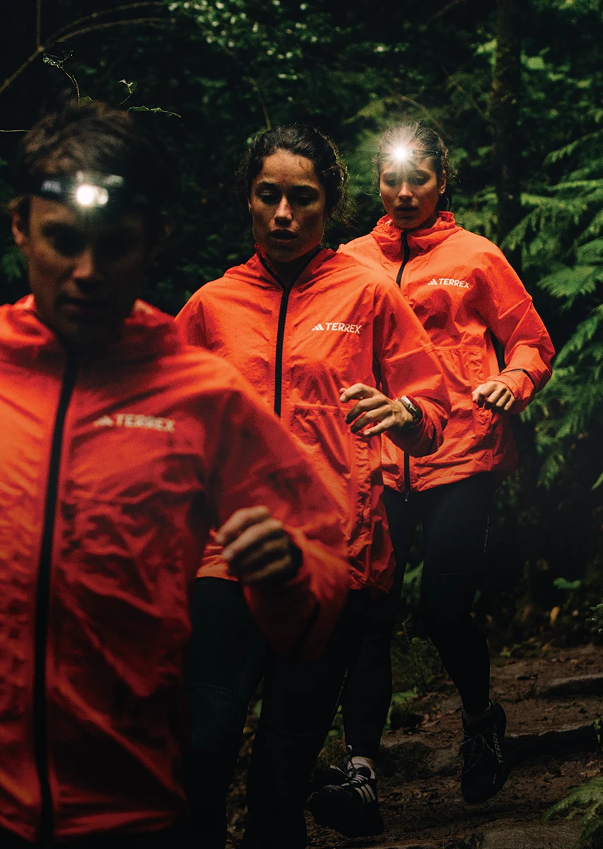 The image shows three individuals wearing orange jackets, likely from a team or company, and they appear to be on a hike or a run in a forested area. They all have headlamp attachments, suggesting they are either out after dark or that it's twilight or overcast.
