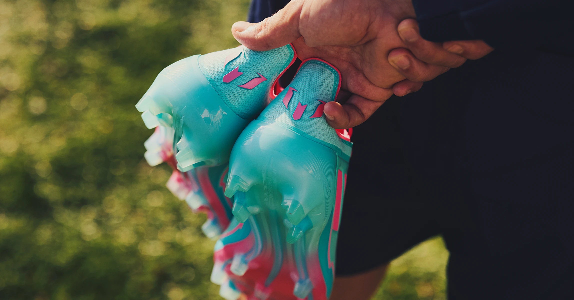 The image shows a person holding up a pair of blue-pink adidas sneakers. The background is out of focus but suggests an outdoor setting.