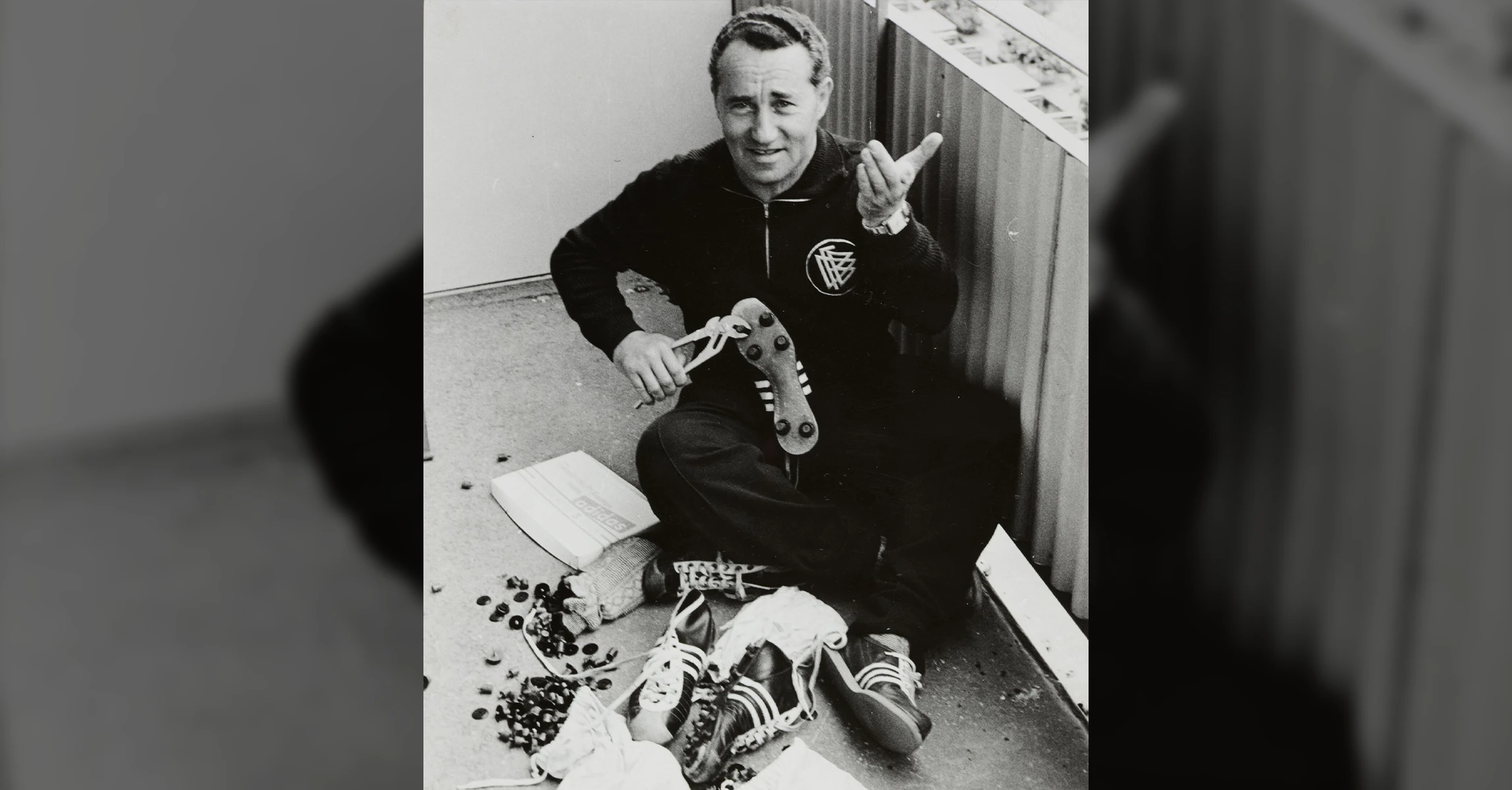 The image shows a person sitting on the floor. It appears to be an old photo, judging by its quality and style of clothing. The person is repairing shoe soles, possibly for football players.