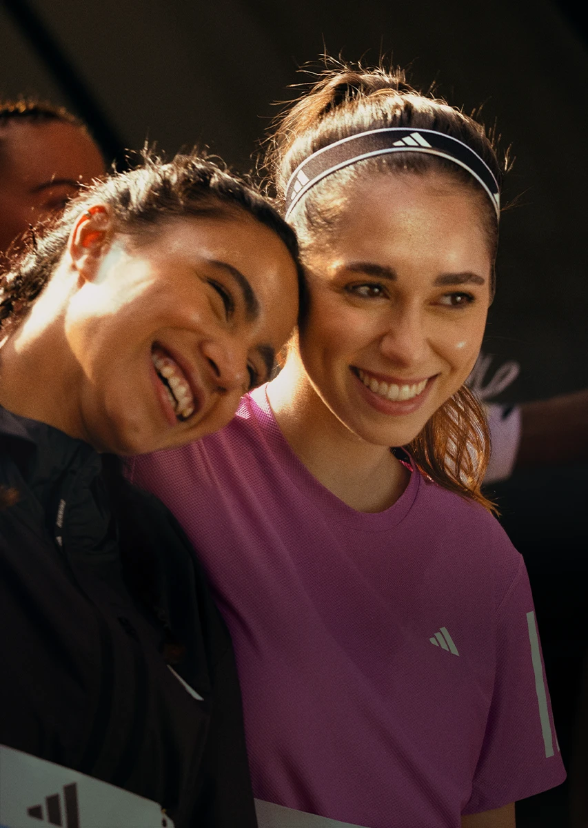 This is a photo featuring two female athletes smiling and looking away from the camera. They appear to be in athletic attire, with one wearing a headband while the other has her hair pulled back into a ponytail. Both are wearing sports jerseys, suggesting they might be part of a team or event related to sports.