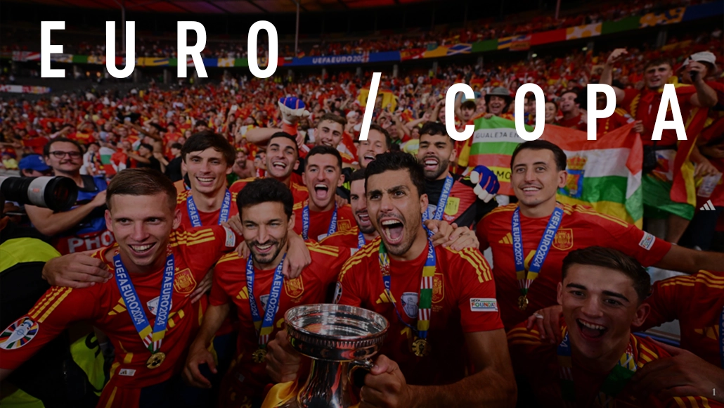 The image shows a team of football players celebrating on what appears to be the pitch after a match. They are wearing red and yellow jerseys, which suggests they could be from a European national team, given the flag in the background. The text "EURO / Copa" indicates that they may have just finished or might be about to start a competition known as EuroCop, an international football tournament for European nations. There is a visible trophy on a podium near them, implying a recent victory or achievement, though no specific details are provided. The team members are expressing joy, with smiles and celebration poses.