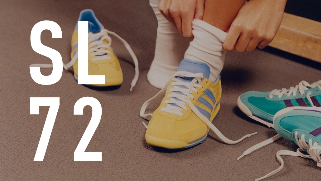 The image shows a pair of yellow sneakers with shoelaces untied, placed on a floor next to other shoes. A person's lower legs are visible above the shoes, wearing white socks.