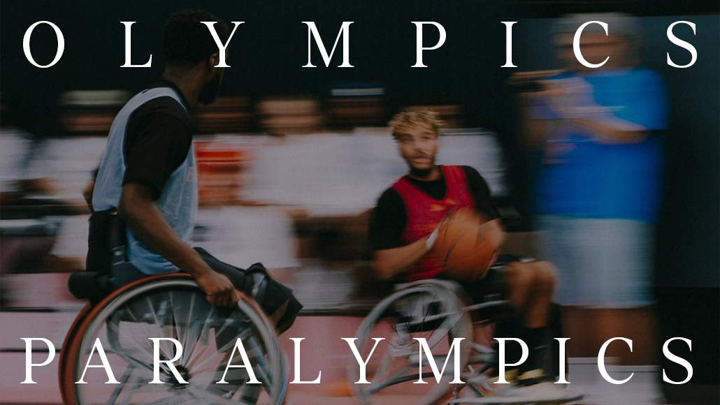 The image displays a person in a wheelchair participating in what appears to be an Olympic games event, where the participant is actively involved and surrounded by people, possibly team members or fellow competitors. The watermark of the Paralympics and text emphasizing this connection is present, indicating that the content focuses on para sports within the Olympic games framework.