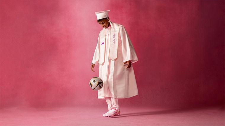 In the image, you see a male model on an advertising or promotional shoot. The model is dressed in vibrant pink, colorful clothing and accessories, usually worn for graduations. He is kicking a football.