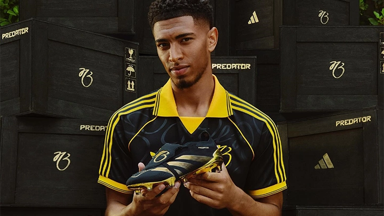 The person in the image appears to be holding a shoe and standing next to cases that likely contain more shoes. His facial expression suggests a positive mood, possibly related to his professional achievement or announcement. The person is dressed in what seems like sports attire, suggesting he may be involved with a sporting event or is in the context of sports. The logo in the background appears to be associated with Adidas and the word "PREDATORS," which might refer to a team name or to the brand itself if it's a product name.