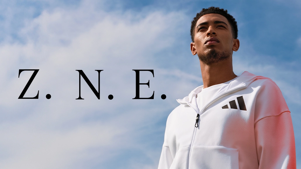 The image shows a person standing outdoors posing for the camera. The individual is wearing a white hooded sweatshirt with an adidas logo visible on it, and there's an outdoor setting with a blue sky backdrop behind them. 