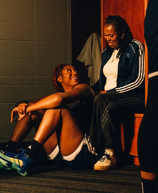 The image shows two women sitting on the floor. One person appears to be a female athlete wearing sports attire, and the other person is an adult female with one arm propped up on her knee. They are both looking at something off-camera with expressions of surprise or amazement.