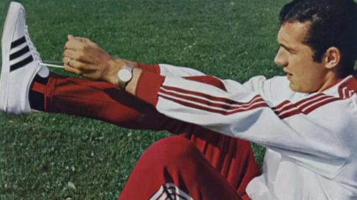 The person shown in the image appears to be putting on his training shoes during an outdoors training session, as indicated by the grass turf of a soccer field.