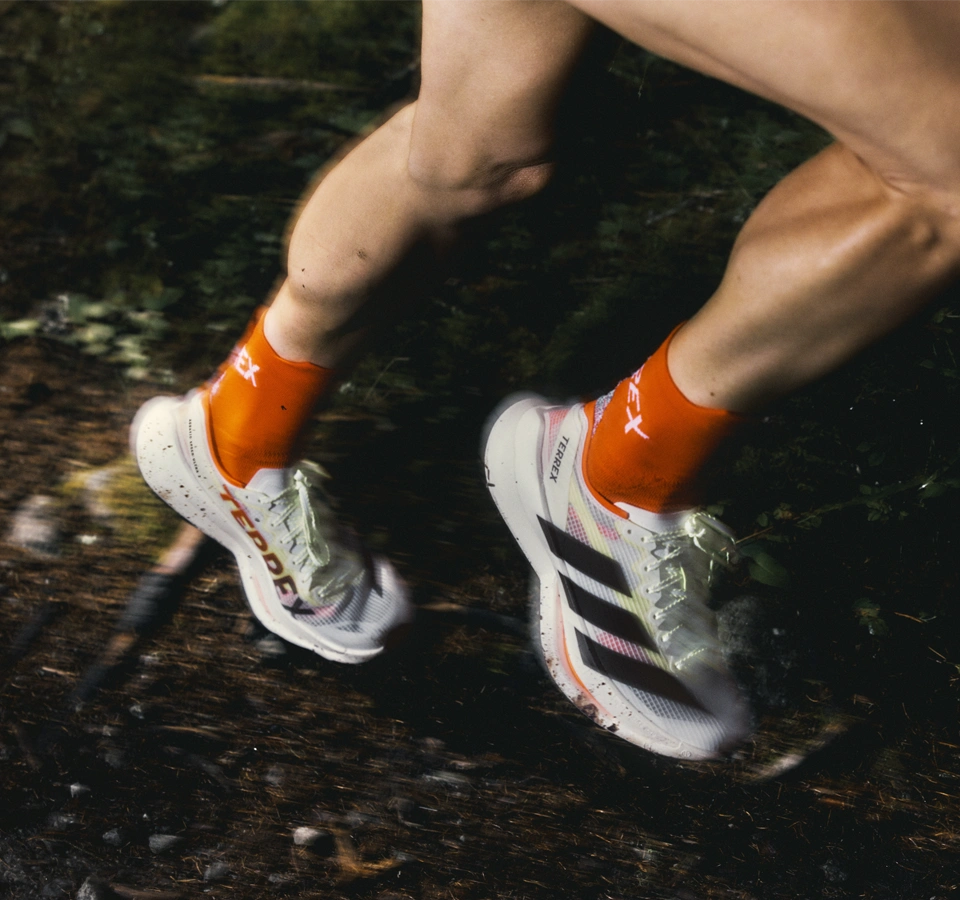 Laufende Person mit weißen adidas trailrunning Schuhen und orangefarbenen Socken auf einem Waldweg. (Foto)