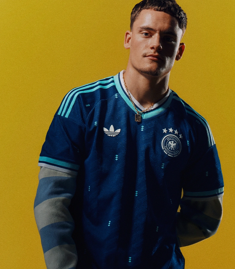 Jude Bellingham in weißem Tanktop und schwarzer adidas Trainingshose blickt lächelnd zu einem schwebenden Fußball vor einem rot-grünen Hintergrund. (Foto)