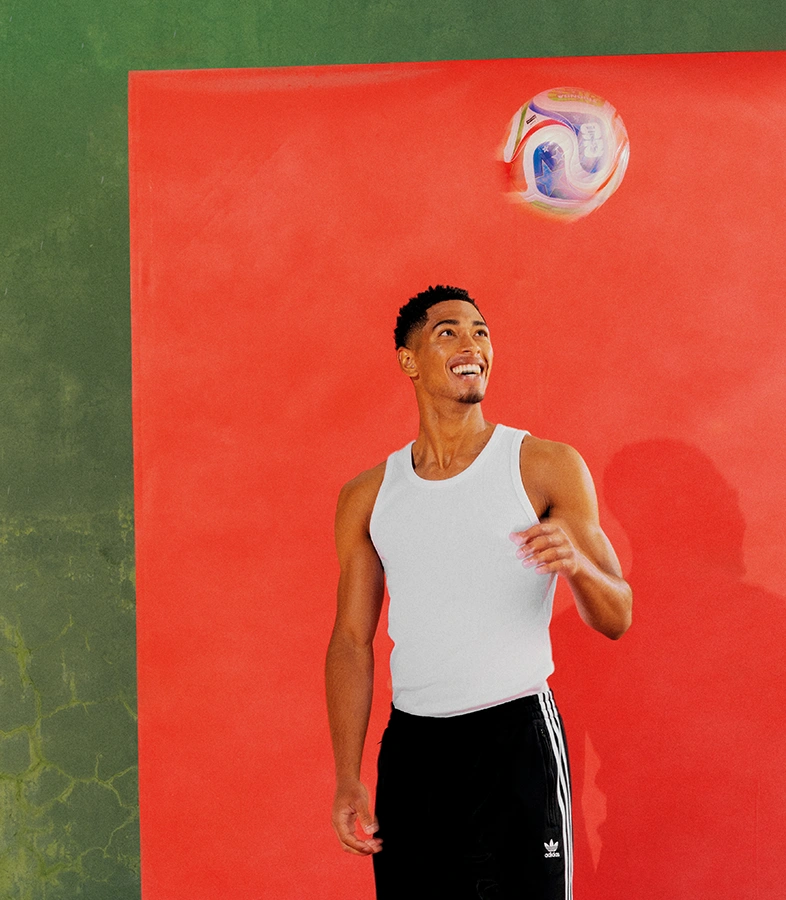 Jude Bellingham in weißem Tanktop und schwarzer adidas Trainingshose blickt lächelnd zu einem schwebenden Fußball vor einem rot-grünen Hintergrund. (Foto)
