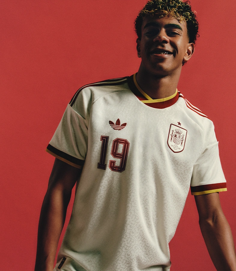 Lamine Yamal trägt ein ärmelloses adidas Shirt, Jeansshorts und grüne adidas Sneaker und spielt mit einem Fußball vor einer gelb-grauen Wand. (Foto)