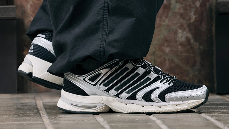 Nahaufnahme von schwarzen und silbernen Sneakern an einer Person in weiter schwarzer Hose, stehend auf einem Steinboden. (Foto)