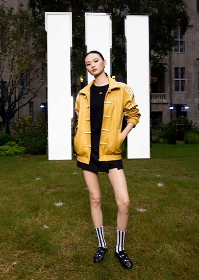 Model wearing a yellow adidas jacket standing on grass in front of three white stripes. (Photo)