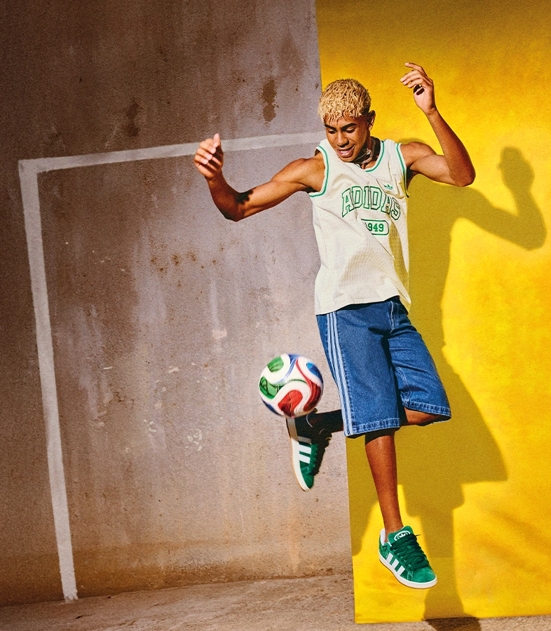 Lamine Yamal wearing a sleeveless adidas shirt, denim shorts and green adidas sneakers plays with a football against a yellow and grey wall. (Photo)